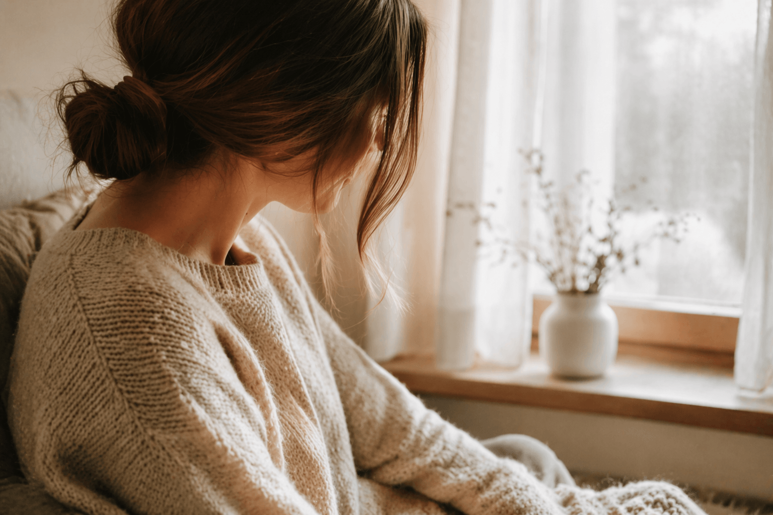 Frau mit lockerem Dutt sitzt in beigem Strickpullover am Fenster in einer ruhigen, warmen Wohnumgebung.