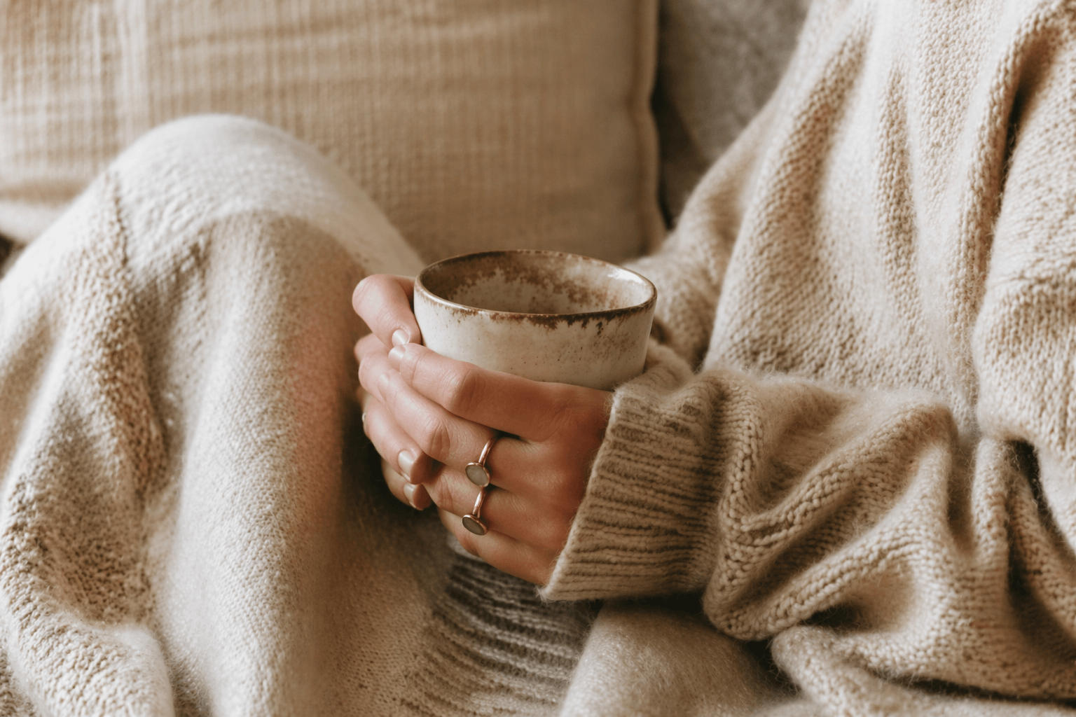 Hände einer Frau in beigem Strickpullover halten eine Keramiktasse in einer ruhigen, warmen Wohnatmosphäre.