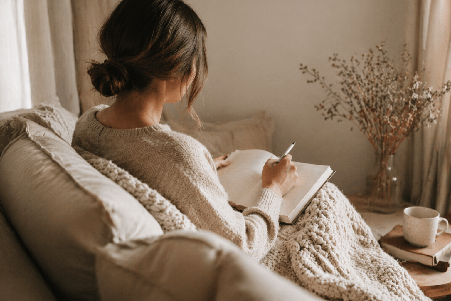 Frau sitzt eingekuschelt in einem Sessel und schreibt in ein Notizbuch in einer warmen, gemütlichen Ecke.