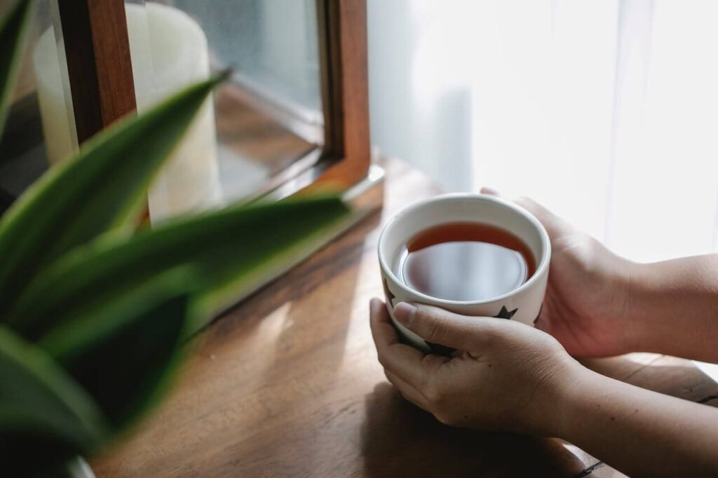 Hände halten morgens eine warme Tasse Tee am Fenster im sanften Tageslicht