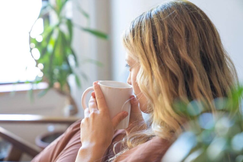Frau sitzt mit einer Tasse am Fenster im Morgenlicht und genießt einen ruhigen Moment für mehr Energie und ein entspannteres Nervensystem.