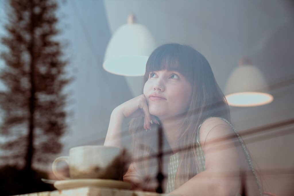 Nachdenkliche Frau sitzt mit einer Tasse Kaffee am Fenster und wirkt mental erschöpft und in Gedanken versunken.