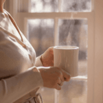 Frau mit Tasse am Fenster am Morgen - Müdigkeit und sanfte Morgenroutine für mehr Energie im Mama-Alltag