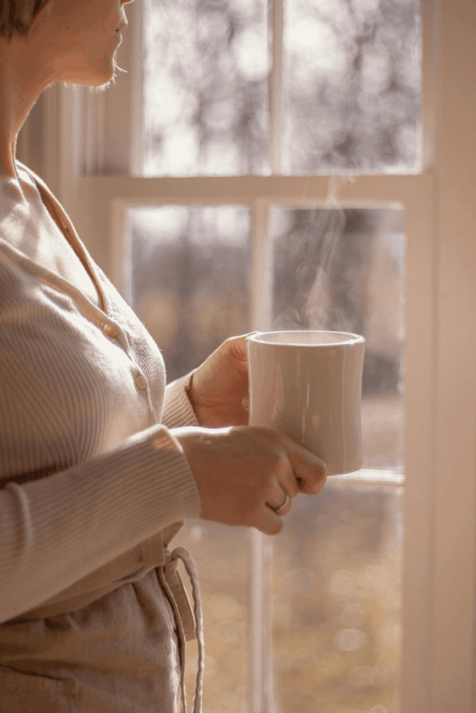 Frau mit Tasse am Fenster am Morgen - Müdigkeit und sanfte Morgenroutine für mehr Energie im Mama-Alltag
