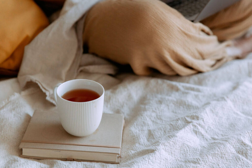 Tasse Tee auf einem Buch in einem gemütlichen Bett mit heller Bettwäsche.