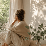 Frau sitzt nachdenklich am Fenster in einem hellen, ruhigen Zuhause. Warmes Morgenlicht fällt durch die Vorhänge und schafft eine entspannte Atmosphäre. Das Bild steht sinnbildlich für ein überreiztes Nervensystem, innere Anspannung und den Wunsch nach mehr Ruhe im Mama-Alltag.
